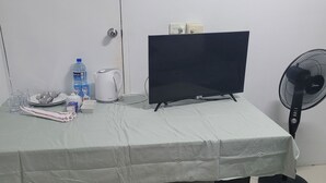 A small, neatly arranged room with a bed, a table with a TV, a kettle, and a fan.