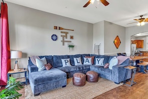 A living room with a blue sofa, leather ottomans, a coffee table, a red curtain, a lamp, and a wall with decorative items including a 'PLAY' sign.
