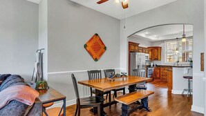 A spacious dining area with a wooden table and chairs, a ceiling fan, and a kitchen in the background. - Stay & Play! Right next to SDC + Waffles, Games, Hottub, Pool, Outdoor Movies! (Branson)