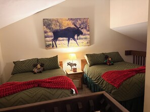 Ein Schlafzimmer mit zwei Betten, einem Nachttisch mit Lampe, einem Elchgemälde an der Wand und einer Stoffelch auf einem Bett.