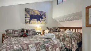 Une chambre à coucher avec deux lits, chacun recouvert d’une courtepointe à motifs d’élans, une table de chevet avec une lampe et un tableau représentant un élan, accroché au mur.