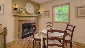 A dining room with a brick fireplace, wooden chairs, a table with a floral tablecloth, and a window with a view of greenery.