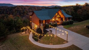 A log cabin with a porch, surrounded by a string of lights, set against a forest backdrop. - Dancing Bear Lodge – Hear the Dollywood Train & Soak in Unforgettable Smoky Mountain Views (Sevierville)