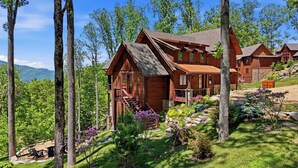 A wooden cabin with a stone foundation, surrounded by lush greenery and trees. - 3BR LUX Cabin w Firepit, Game Loft + Scenic Views! (Banner Elk)