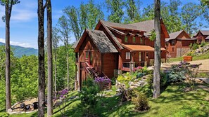 A wooden cabin with a stone foundation, surrounded by lush greenery and trees. - 3BR LUX Cabin w Firepit, Game Loft + Scenic Views! (Banner Elk)