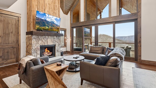 A spacious living room with a stone fireplace, leather sofas, and a large TV. - 3BR LUX Cabin w Firepit, Game Loft + Scenic Views! (Banner Elk)