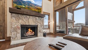 A cozy living room with a stone fireplace, a leather sofa, and a large window overlooking a mountainous landscape. - 3BR LUX Cabin w Firepit, Game Loft + Scenic Views! (Banner Elk)
