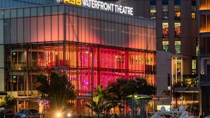 Ein modernes Gebäude mit einer großen Glasfassade, einem gut sichtbaren Schild mit der Aufschrift 'ASB Waterfront Theatre' und einem Spiegelbild im Wasser davor.