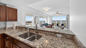 A kitchen with granite countertops, stainless steel sink, and wooden cabinets. There is a balcony with a view of the ocean and a ceiling fan. - Endless Horizon at the Aruba (Daytona Beach)