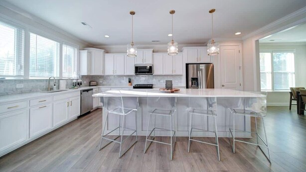 A modern kitchen with white cabinets, a large island with bar stools, stainless steel appliances, and pendant lighting.