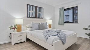 Une chambre moderne avec un lit blanc, deux tables de chevet avec des lampes, une fenêtre avec des rideaux et une œuvre d’art abstraite au mur.