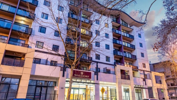 Un immeuble à plusieurs étages, sur lequel on peut voir, au niveau du rez-de-chaussée, une grande enseigne indiquant le nom « WALDORF », ce qui suggère qu’il s’agit d’un hôtel ou d’un établissement similaire.