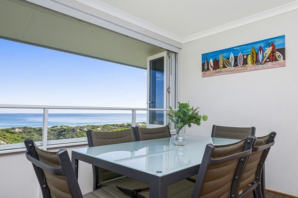 A dining area with a glass table and chairs, a vase with greenery, and a painting of surfboards on the wall.
