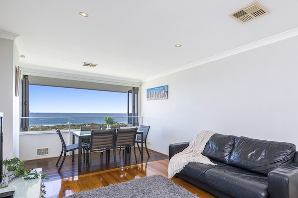 A modern living room with a view of the ocean, a dining area with chairs, and a black leather sofa. - Renovated Ocean-View Scarborough Apartment | Walk to Beach & Amphitheatre (Scarborough)