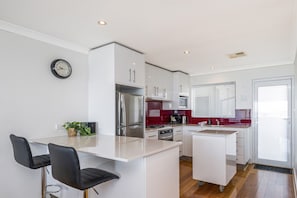 A modern kitchen with white cabinetry, a stainless steel refrigerator, a dining area with two black chairs, and a red backsplash. - Renovated Ocean-View Scarborough Apartment | Walk to Beach & Amphitheatre (Scarborough)