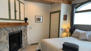 Un dormitorio con chimenea de piedra, una cama con ropa de cama a rayas, una lámpara de noche y vistas al exterior a través de una ventana.