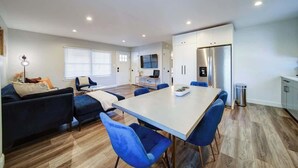 A modern living room with a sofa, a dining table with chairs, a TV, and a kitchen area with cabinets and appliances.