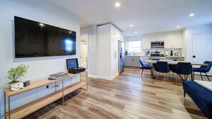 A modern living room with a flat-screen TV, wooden console, and a dining area with blue chairs.