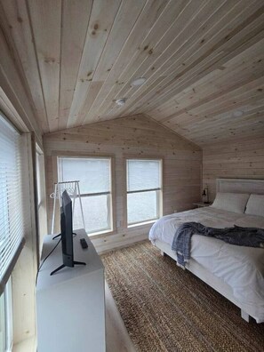 A bedroom with a sloped ceiling, two windows with blinds, a bed with white bedding, a bedside table with a lamp, and a television on a stand.
