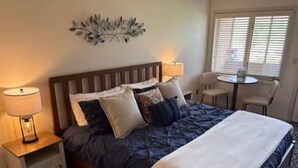 A neatly made bed with a blue quilt, white pillows, and a white throw blanket. There is a wooden headboard, a small bedside table with a lamp, and a chair next to a window with blinds. - The Grand Duplex of Silvercreek Inn. Combination of two lock off units. (Granby)