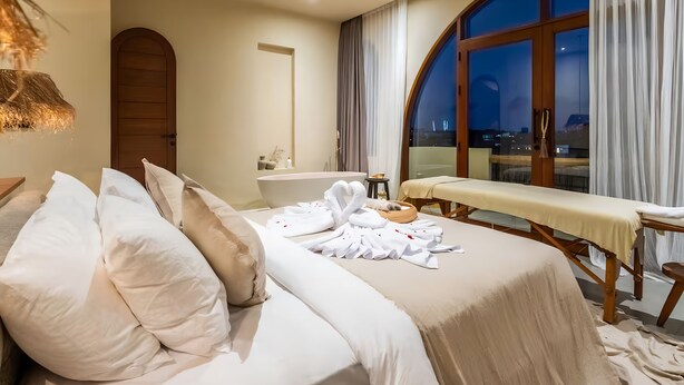 Un dormitorio moderno con una cama grande, una mesa de masajes, una bañera y vistas de la ciudad al anochecer.
