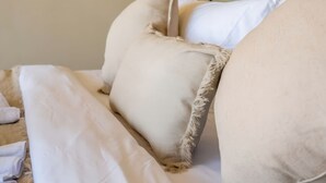Una cama con ropa de cama blanca y una almohada color beige.