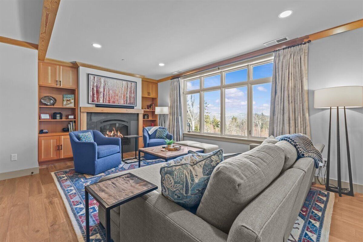 A living room with a fireplace, blue armchair, grey sofa, and a large window with curtains.