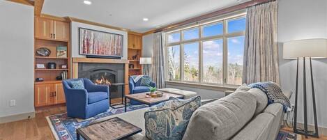 A living room with a fireplace, blue armchair, grey sofa, and a large window with curtains.