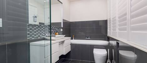 A modern bathroom with a glass shower enclosure, a white sink, a bathtub, and two toilets.