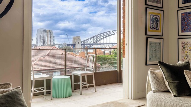 Un balcón con vista al Puente de Sydney, una silla blanca, una mesita redonda y un sofá con cojines.