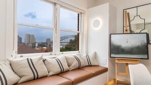Una habitación con asiento junto a la ventana, un cuadro sobre caballete y vistas a un paisaje urbano que incluyen un puente.