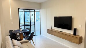 A modern living room with a large flat-screen TV, a sofa, a dining table, and a sliding glass door leading to a balcony. - Apart Hotel recém reformado TV de cinema (Salvador)
