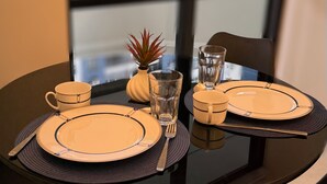 A dining table set with plates, cutlery, and a glass of water, with a view of buildings through a window. - Apart Hotel recém reformado TV de cinema (Salvador)