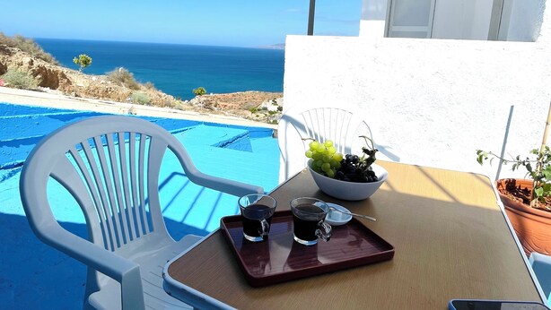 A white chair and table with a tray of coffee and grapes, overlooking a blue pool and the sea.