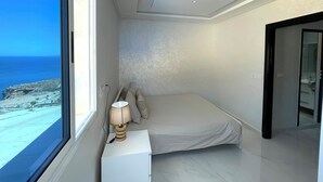 A bedroom with a bed, a nightstand with a lamp, and a view of the sea through a large window.