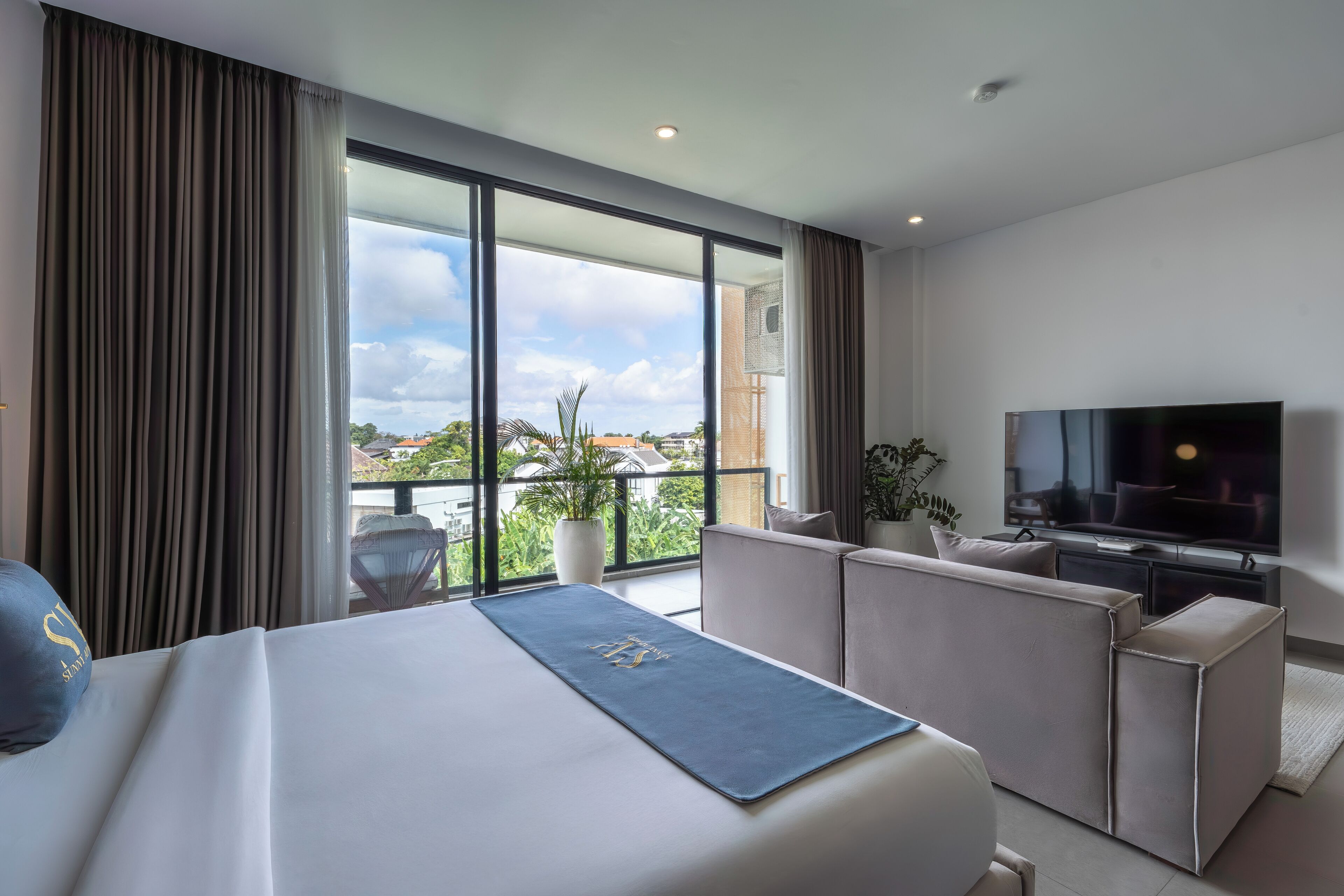Una habitación de hotel moderna con una cama grande, un televisor de pantalla plana, un balcón con vistas a la ciudad y una zona de estar con una planta.