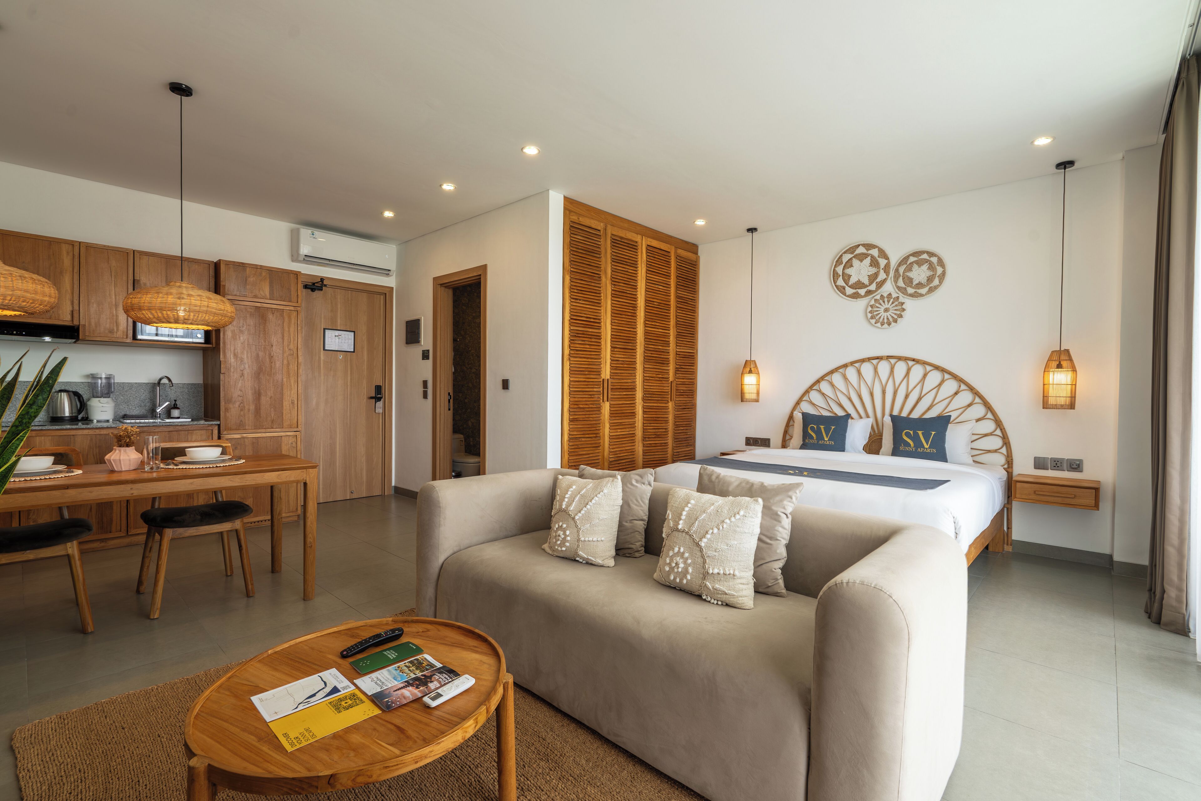 A modern hotel room with a sofa, a bed with decorative pillows, a wooden table, and a kitchen area.