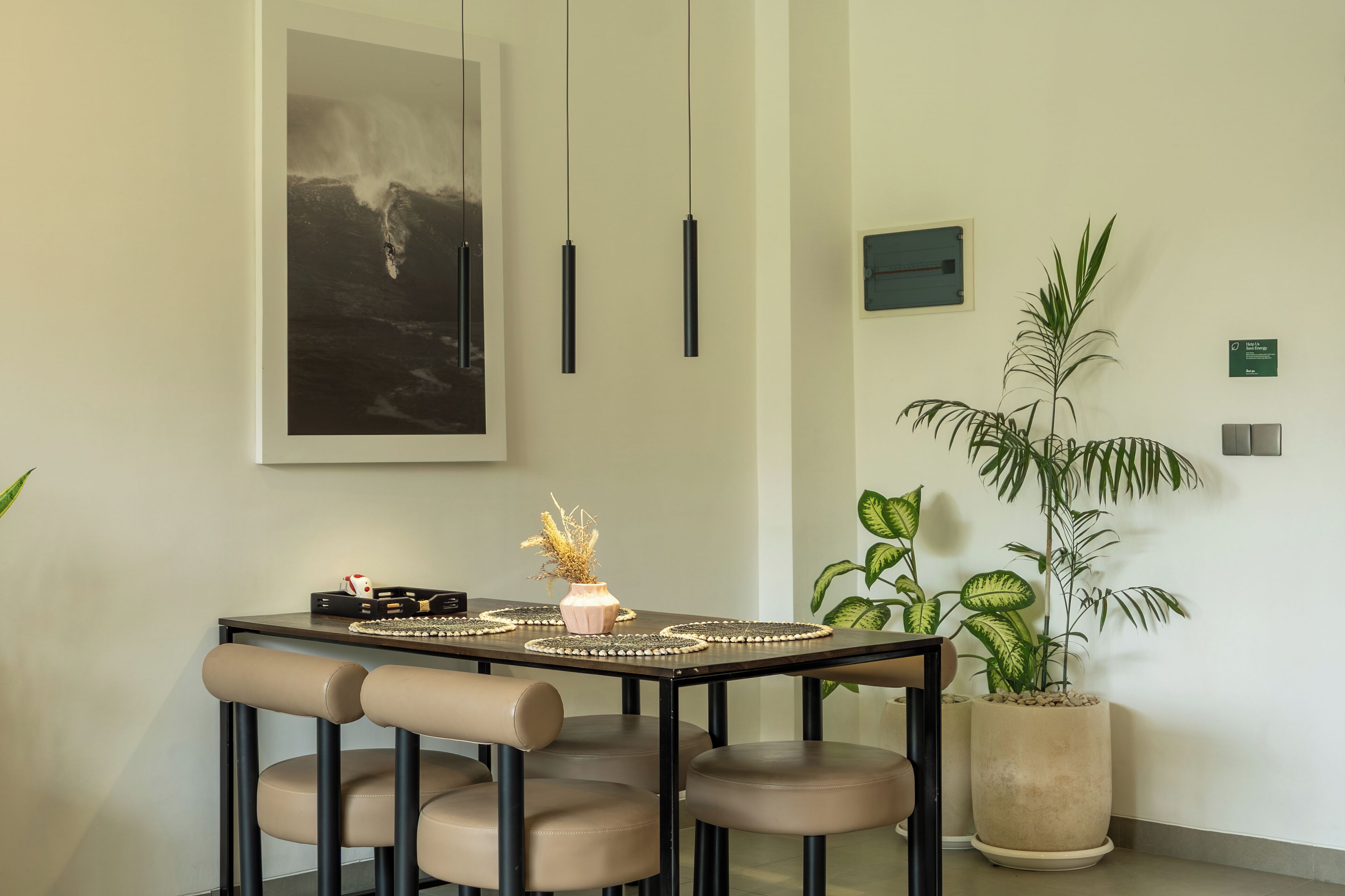 A dining area with a table set for four, a large framed artwork above, pendant lights, and potted plants.