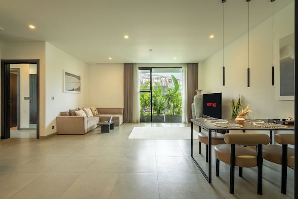 A modern living room with a sofa, a dining table with chairs, a TV, and a view of greenery outside. - Sunny Aparts 1 by Betterplace (Tibubeneng)