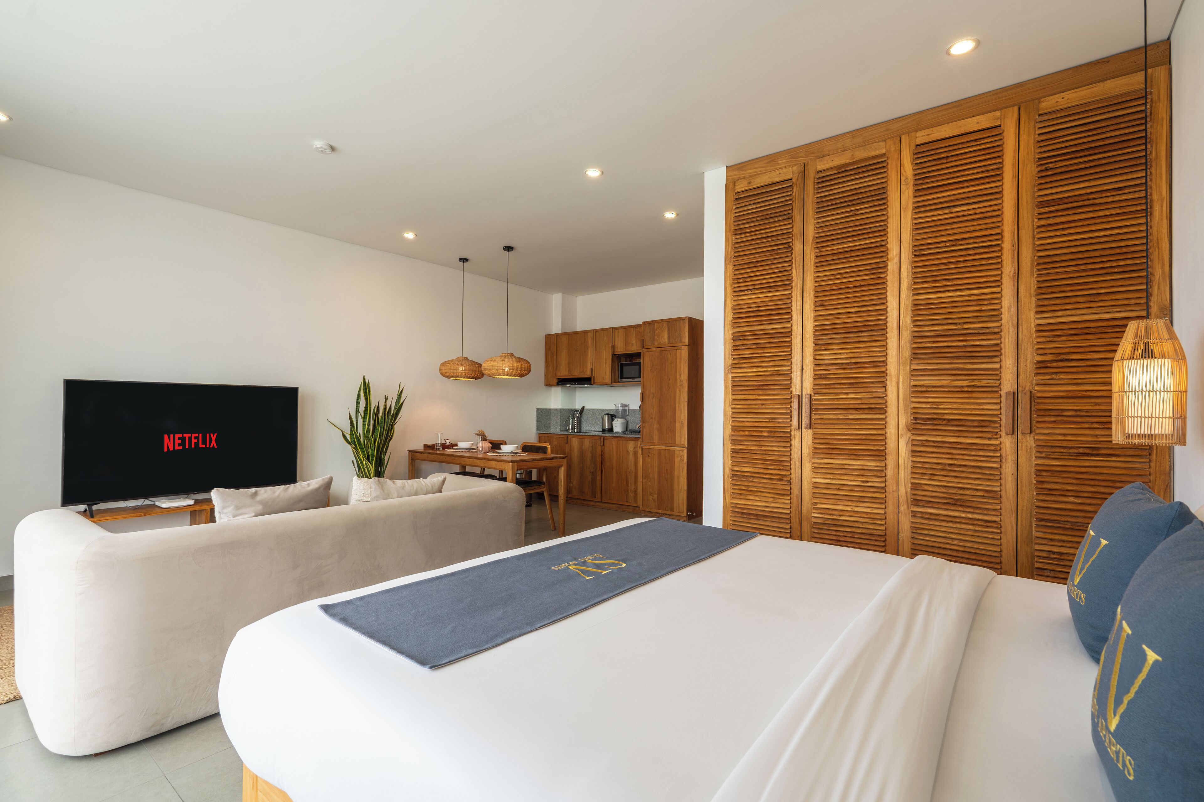 Ein modernes Hotelzimmer mit einem großen Bett, einer Couch, einem Fernseher mit 'NETFLIX' und einer Küchenzeile sowie einem Holzschrank.