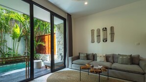 A modern living room with a large glass door leading to a pool, a grey sofa with decorative pillows, and a wooden coffee table. - Sunny Aparts 1 by Betterplace (Tibubeneng)