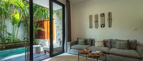 A modern living room with a large glass door leading to a pool, a grey sofa with decorative pillows, and a wooden coffee table.