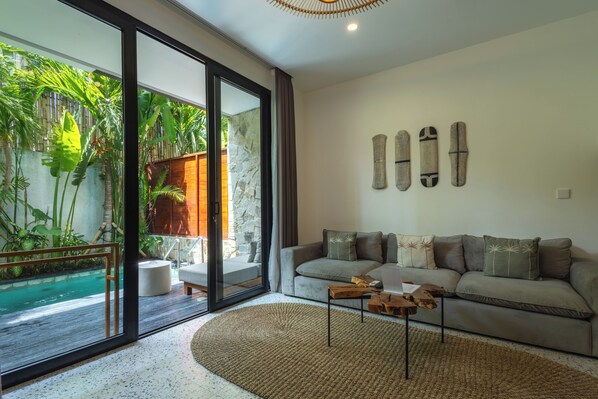 A modern living room with a large glass door leading to a pool, a grey sofa with decorative pillows, and a wooden coffee table. - Sunny Aparts 1 by Betterplace (Tibubeneng)