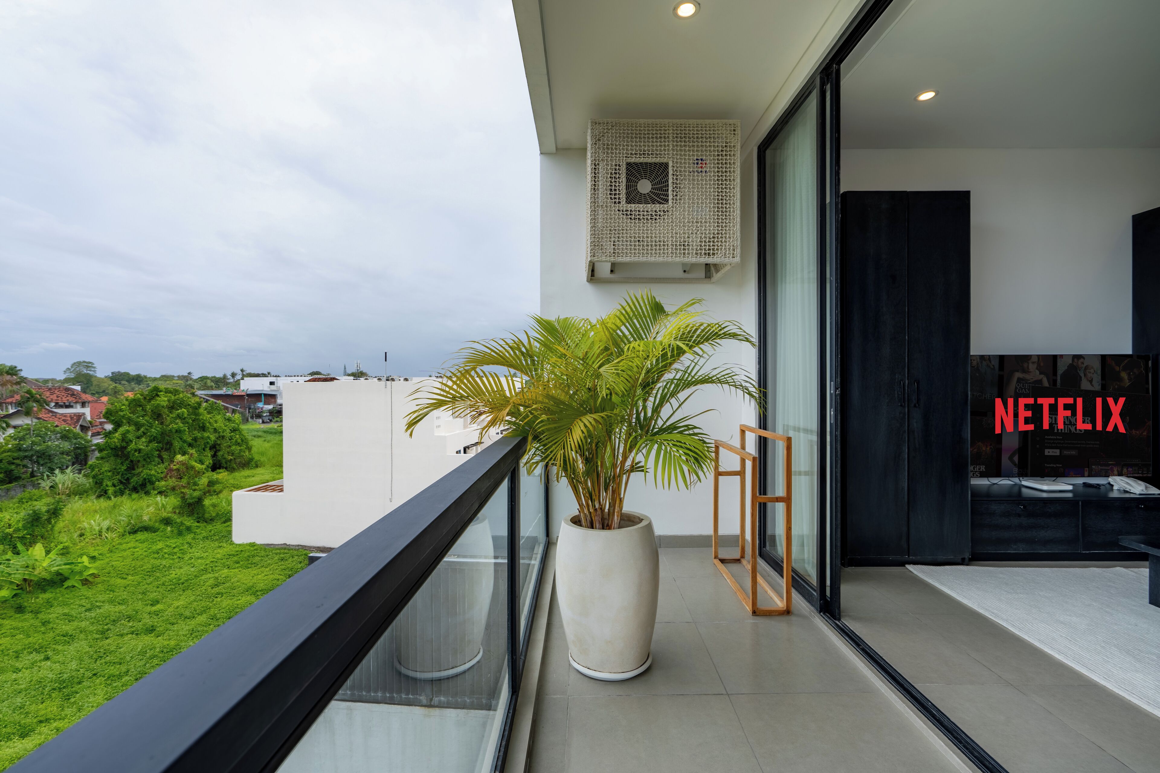 Un balcón con una planta en maceta, un ventanal de vidrio que da a una habitación con el logo de Netflix, y vistas a zonas verdes y edificios.