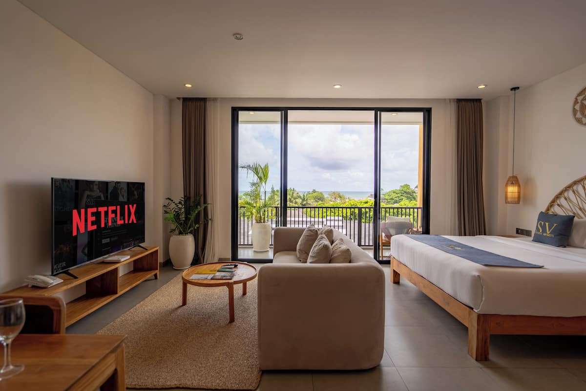 A modern hotel room with a large TV, a sofa, a bed, and a balcony with a view.