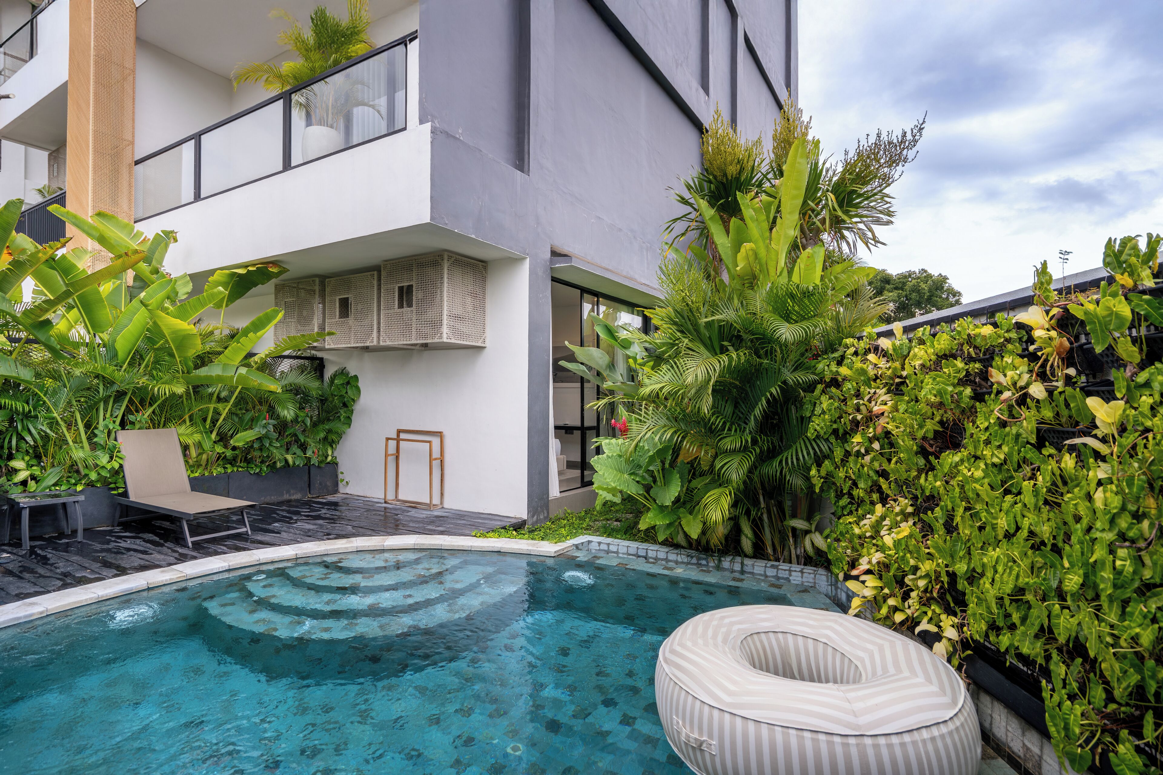 A modern outdoor pool area with a lounge chair and a floating ring, surrounded by lush greenery and a multi-story building with balconies.