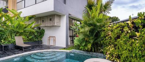 A modern outdoor pool area with a lounge chair and a floating ring, surrounded by lush greenery and a multi-story building with balconies.