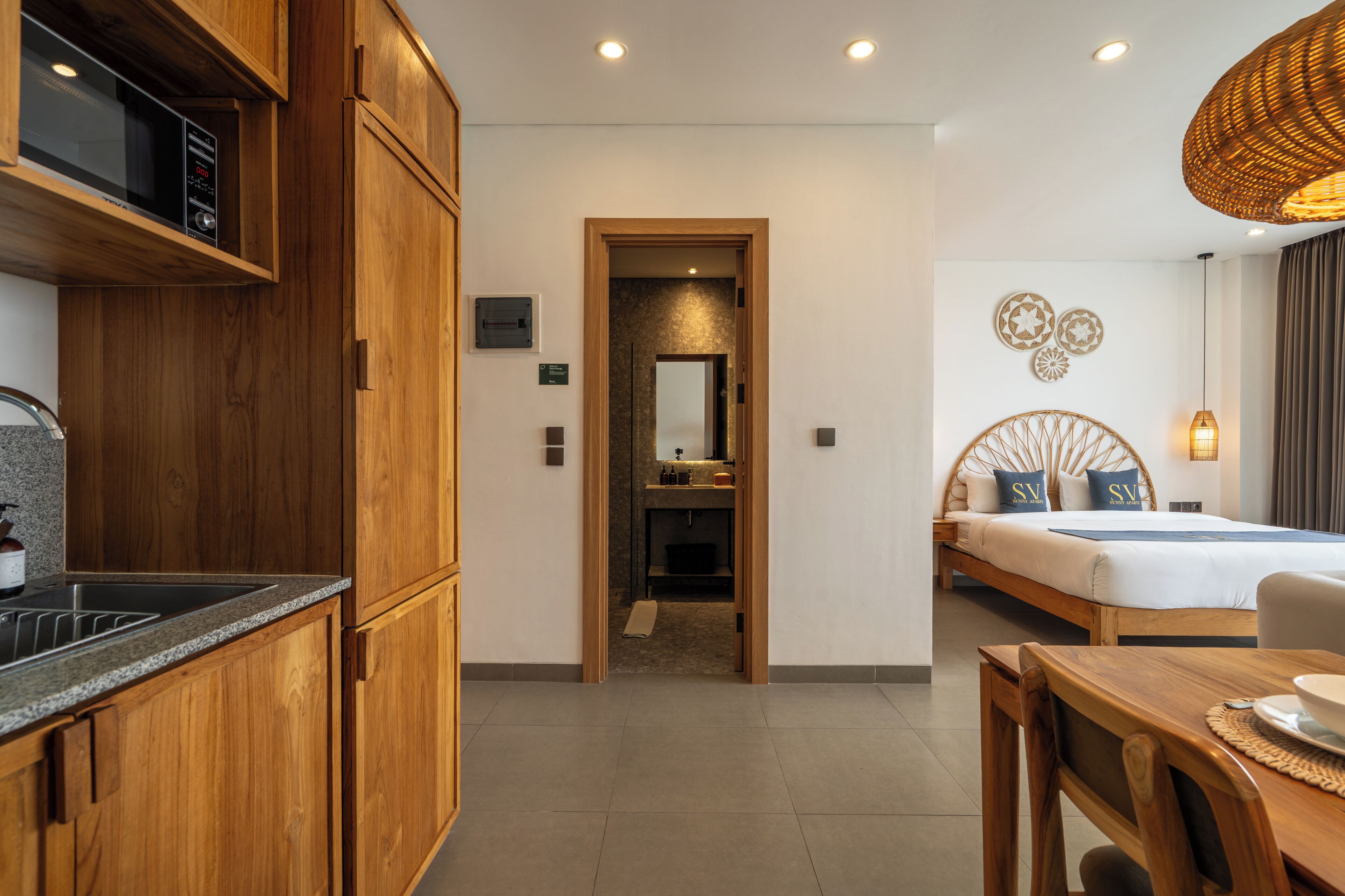 Quarto de hotel moderno com armários de madeira, cozinha compacta, banheiro com espelho e cama com almofadas decorativas.