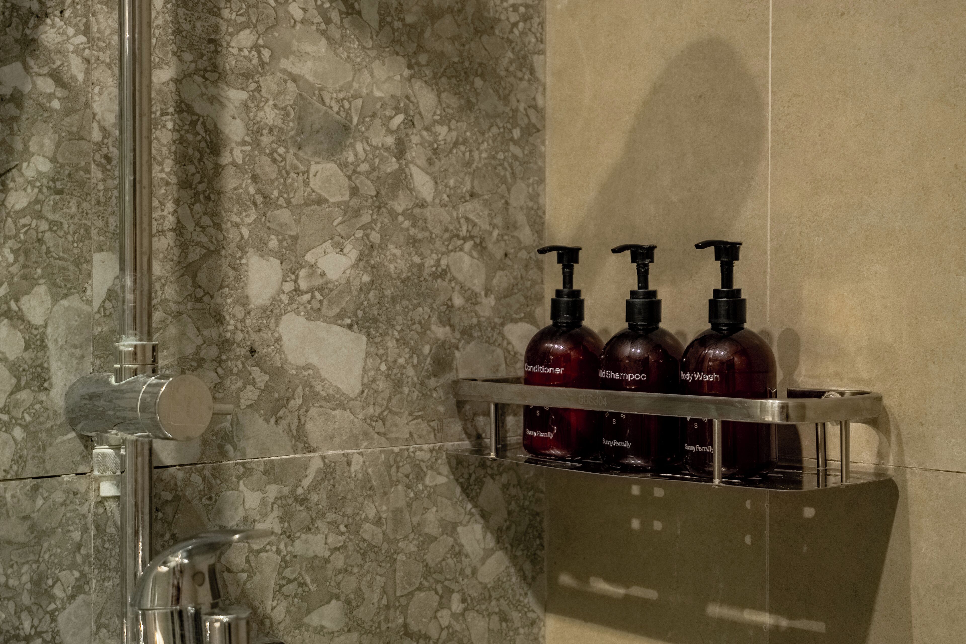 A bathroom with a marble wall, a chrome faucet, and a shelf holding three bottles of shampoo and conditioner.