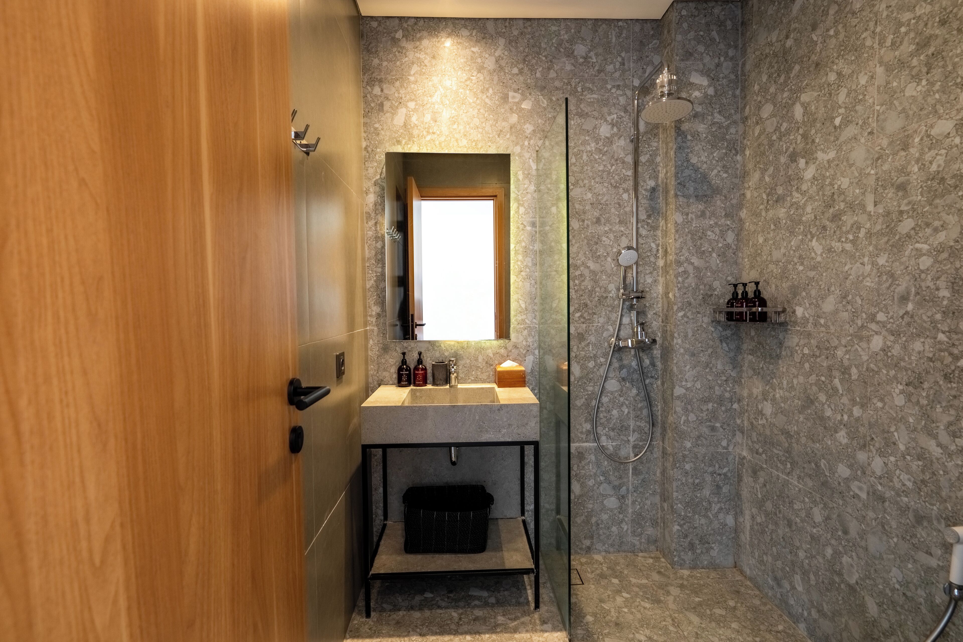 Un baño moderno con puerta de madera, pared de ducha de mármol, ducha con mampara de vidrio y lavamanos con artículos de aseo.